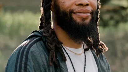 A person with long dreadlocks and a beard is standing outdoors, wearing a jacket over a white shirt, and a necklace with a pendant.
