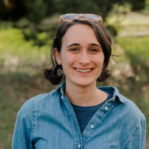 A person in a denim shirt and sunglasses on their head, standing outdoors, smiling at the camera.