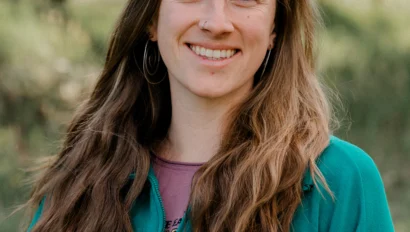 Woman with long brown hair and a teal jacket smiles outdoors with trees in the background.