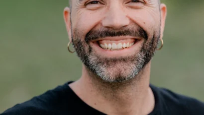 Bald man with a beard and earrings smiling, wearing a black T-shirt that says "Volcom Stone," standing outdoors with a blurred green background.