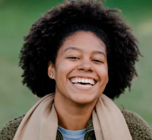Smiling person with curly hair wearing a tan scarf and a green fleece jacket, standing outdoors.