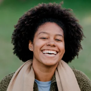 Smiling person with curly hair wearing a tan scarf and a green fleece jacket, standing outdoors.