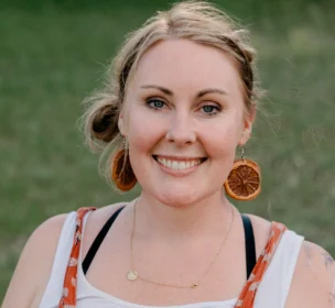 A woman with light hair and orange attire smiles outdoors, wearing large circular earrings.