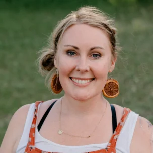 A woman with light hair and orange attire smiles outdoors, wearing large circular earrings.