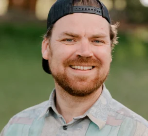 A man with a beard wearing a backward cap and patterned shirt smiles outdoors with a blurred green background.