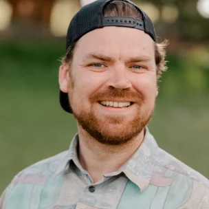 A man with a beard wearing a backward cap and patterned shirt smiles outdoors with a blurred green background.