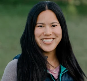 A smiling person with long dark hair wearing a blue vest and gray top stands outdoors.