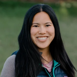 A smiling person with long dark hair wearing a blue vest and gray top stands outdoors.