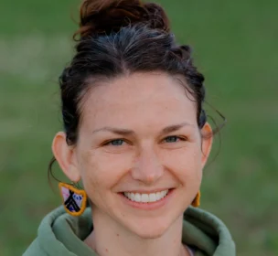 A person with brown hair in a bun, wearing a green hoodie and colorful earrings, smiles in an outdoor setting.