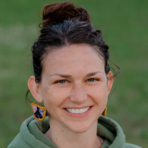 A person with brown hair in a bun, wearing a green hoodie and colorful earrings, smiles in an outdoor setting.