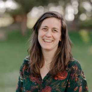 A person with long brown hair, wearing a floral-patterned shirt, smiles outdoors in a grassy area.