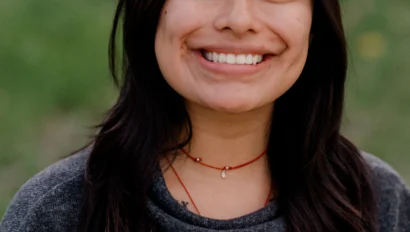 A person with long dark hair and a gray sweatshirt smiles at the camera in an outdoor setting.