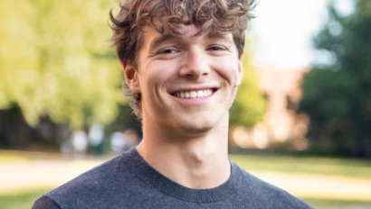 A young man with wavy brown hair and a dark sweater stands outdoors, smiling, with blurred trees and grass in the background.
