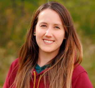 A person with long brown hair and a maroon hoodie smiles outdoors with greenery in the background.