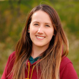 A person with long brown hair and a maroon hoodie smiles outdoors with greenery in the background.