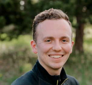 A person in a black jacket smiles outdoors with greenery in the background.