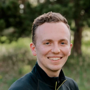 A person in a black jacket smiles outdoors with greenery in the background.