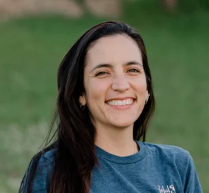 A person with long dark hair wearing a blue t-shirt stands outdoors, smiling. The background is grassy and out of focus.