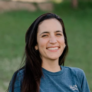 A person with long dark hair wearing a blue t-shirt stands outdoors, smiling. The background is grassy and out of focus.