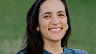 A person with long dark hair wearing a blue t-shirt stands outdoors, smiling. The background is grassy and out of focus.