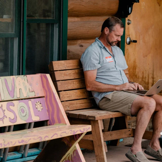 Man sitting on a wooden bench working on a laptop outside a log cabin. A colorful sign reading "Festival del Sol" is beside him.