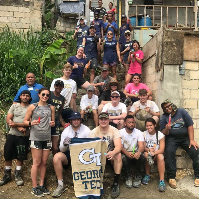 A group of people in casual clothing pose on a muddy slope in front of a building, holding a Georgia Tech banner. Some are wearing gloves and boots, suggesting involvement in a building project.