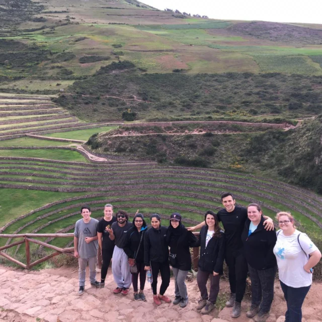 A group of people stand on a stone path with agricultural terraces and green fields in the background.