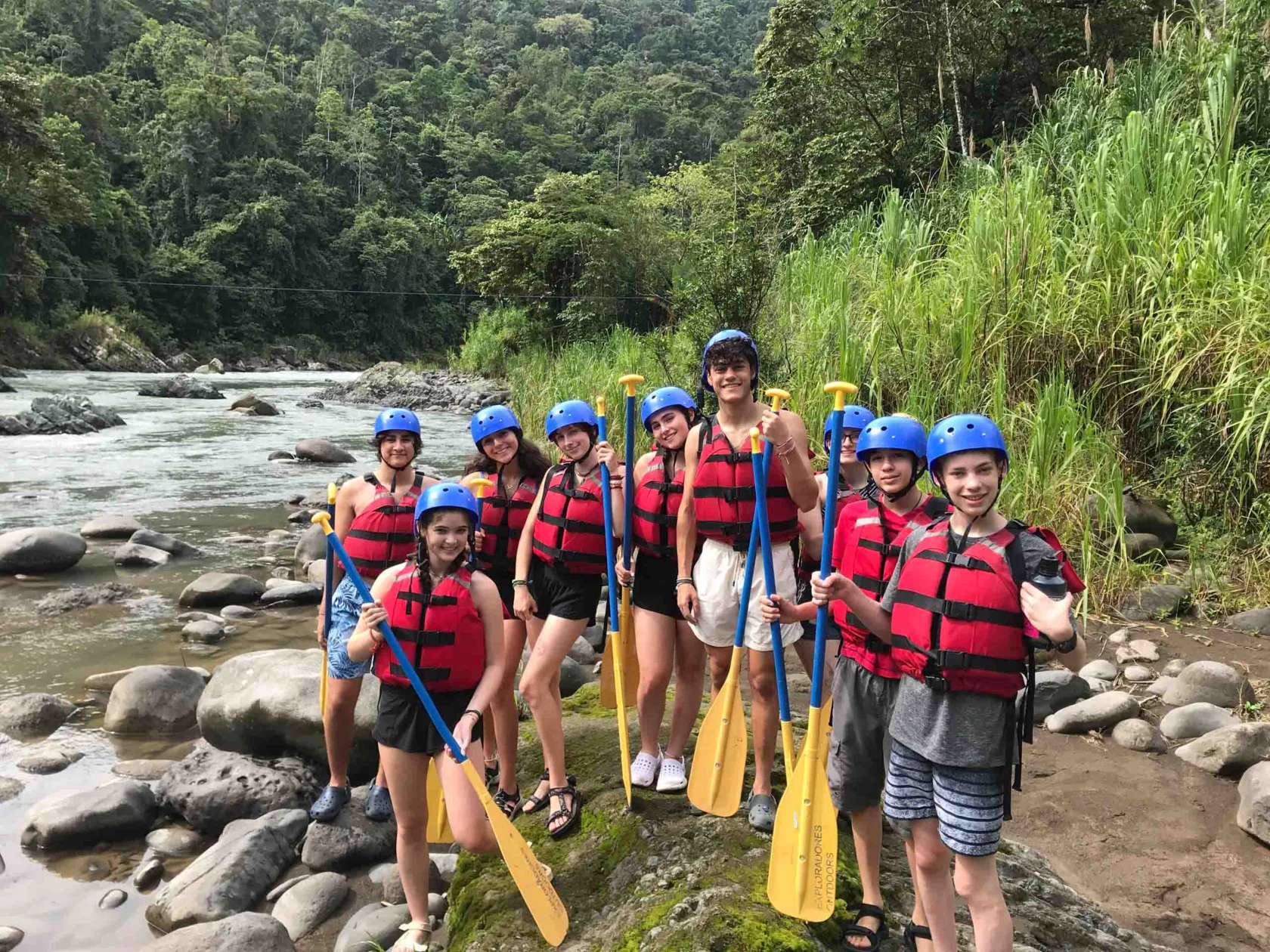 A group of people wearing helmets and life jackets stand by a river holding paddles. Forested hills are in the background.