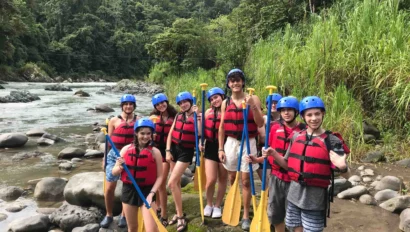 A group of people wearing helmets and life jackets stand by a river holding paddles. Forested hills are in the background.