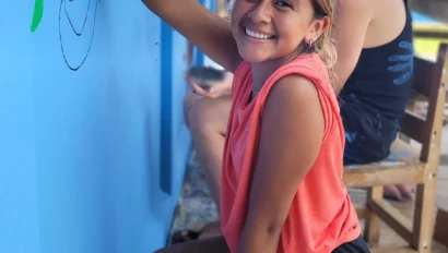 A young girl kneels and smiles while painting a mural on a blue wall. A person is seated behind her, also working on the mural.