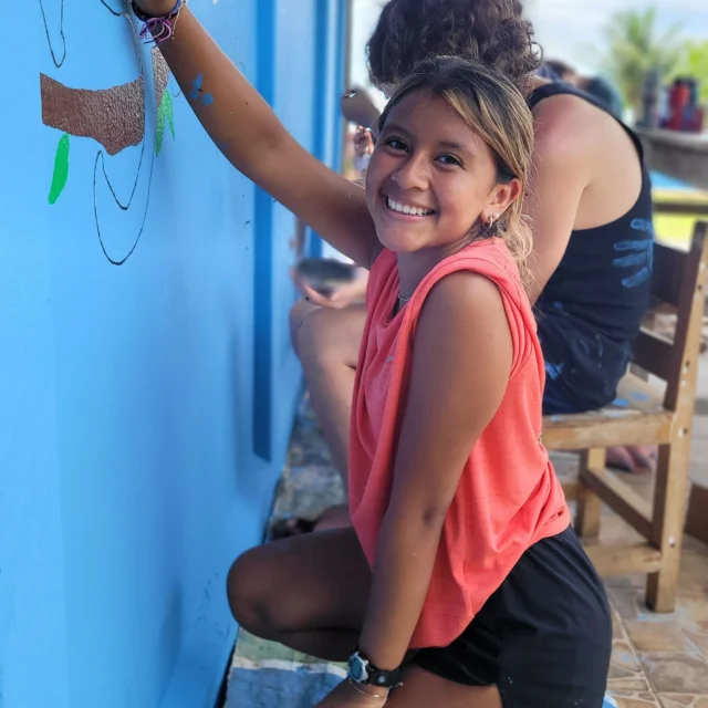 A young girl kneels and smiles while painting a mural on a blue wall. A person is seated behind her, also working on the mural.