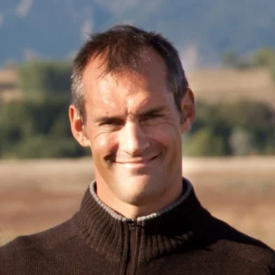 A man in a brown zip-up sweater smiles in an outdoor setting with trees and hills in the background.