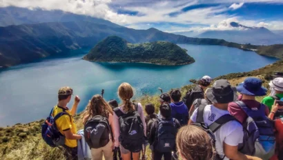 A group of hikers with backpacks stand on a hill overlooking a large lake surrounded by mountains under a partly cloudy sky.