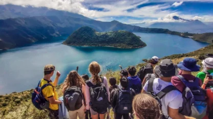 A group of hikers with backpacks stand on a hill overlooking a large lake surrounded by mountains under a partly cloudy sky.