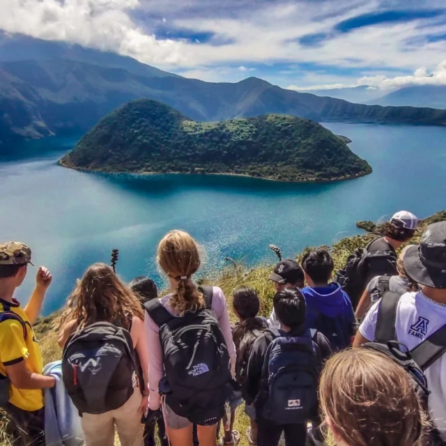 A group of hikers with backpacks stand on a hill overlooking a large lake surrounded by mountains under a partly cloudy sky.