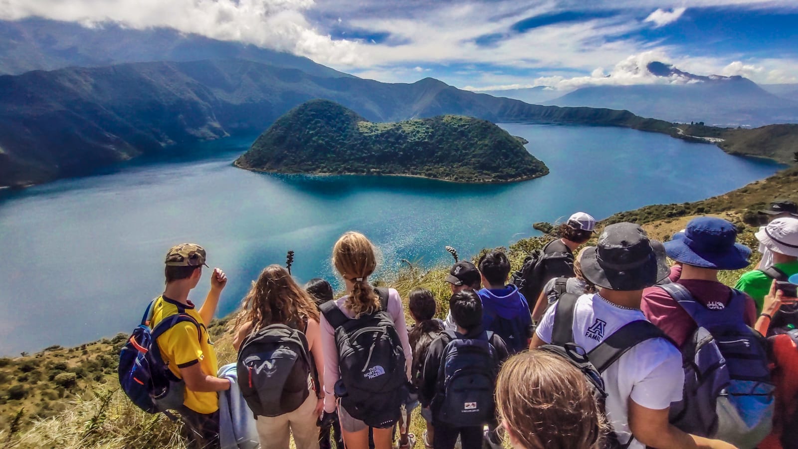 A group of hikers with backpacks stand on a hill overlooking a large lake surrounded by mountains under a partly cloudy sky.