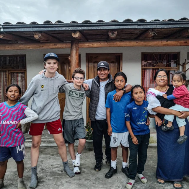 A group of people, including adults and children, stand in front of a building with wooden pillars. Some are wearing casual clothing, and one person is holding a small child.