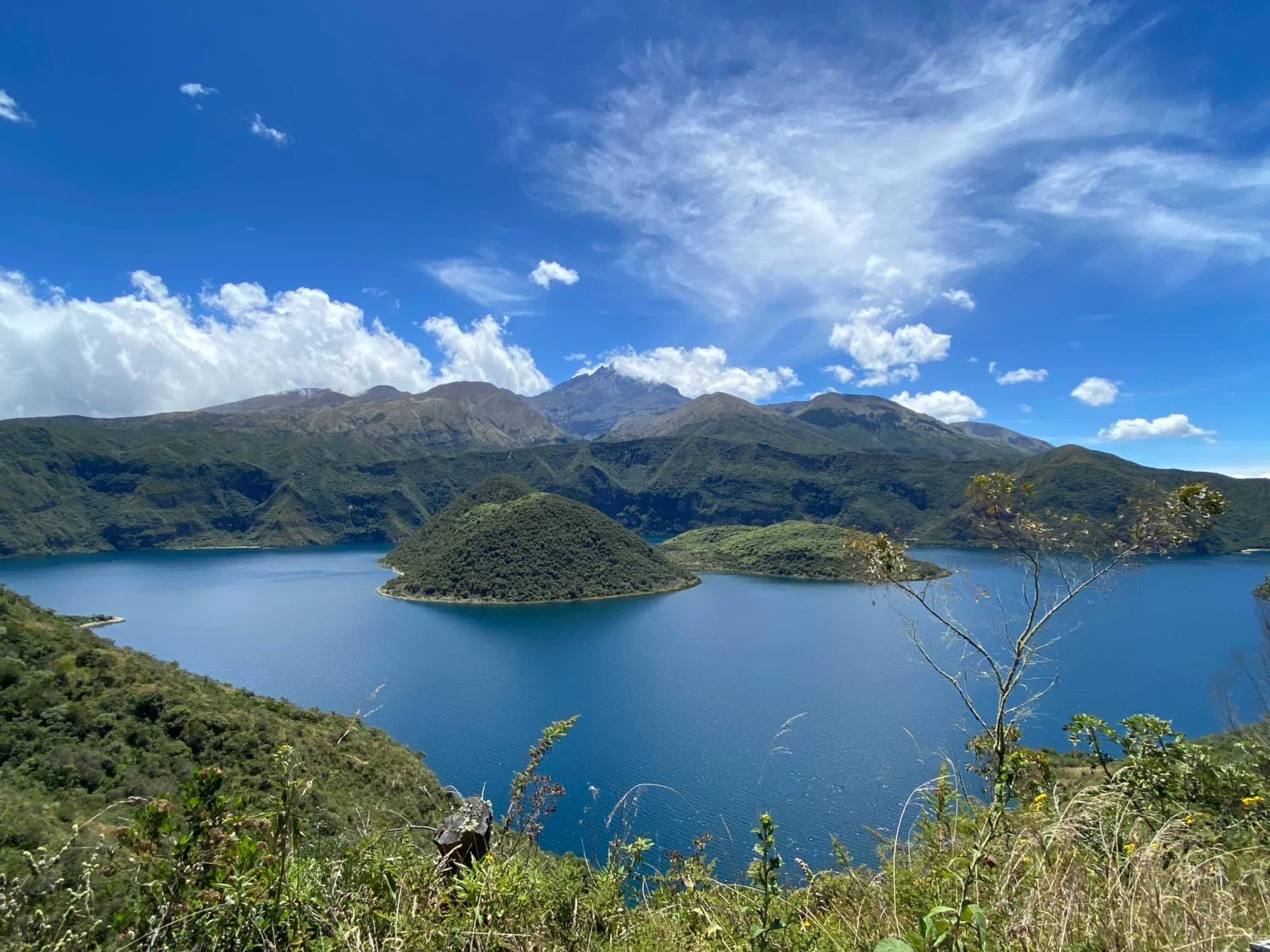 A scenic view of a blue lake surrounded by green hills and mountains under a blue sky with scattered clouds.