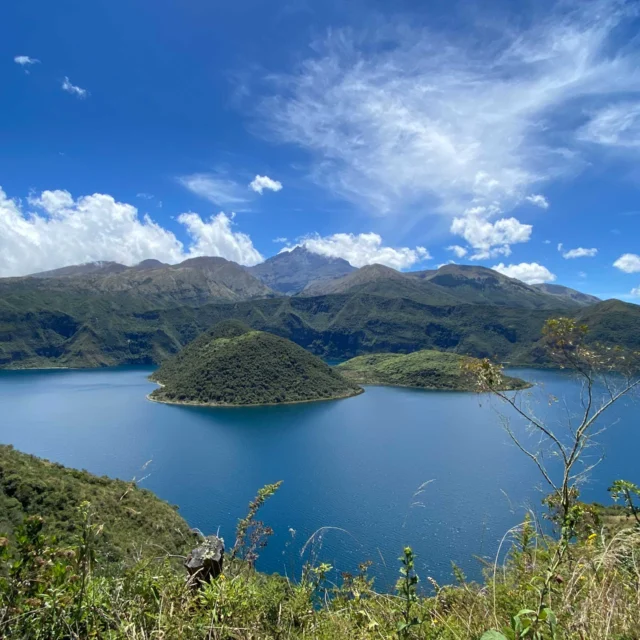 A scenic view of a blue lake surrounded by green hills and mountains under a blue sky with scattered clouds.