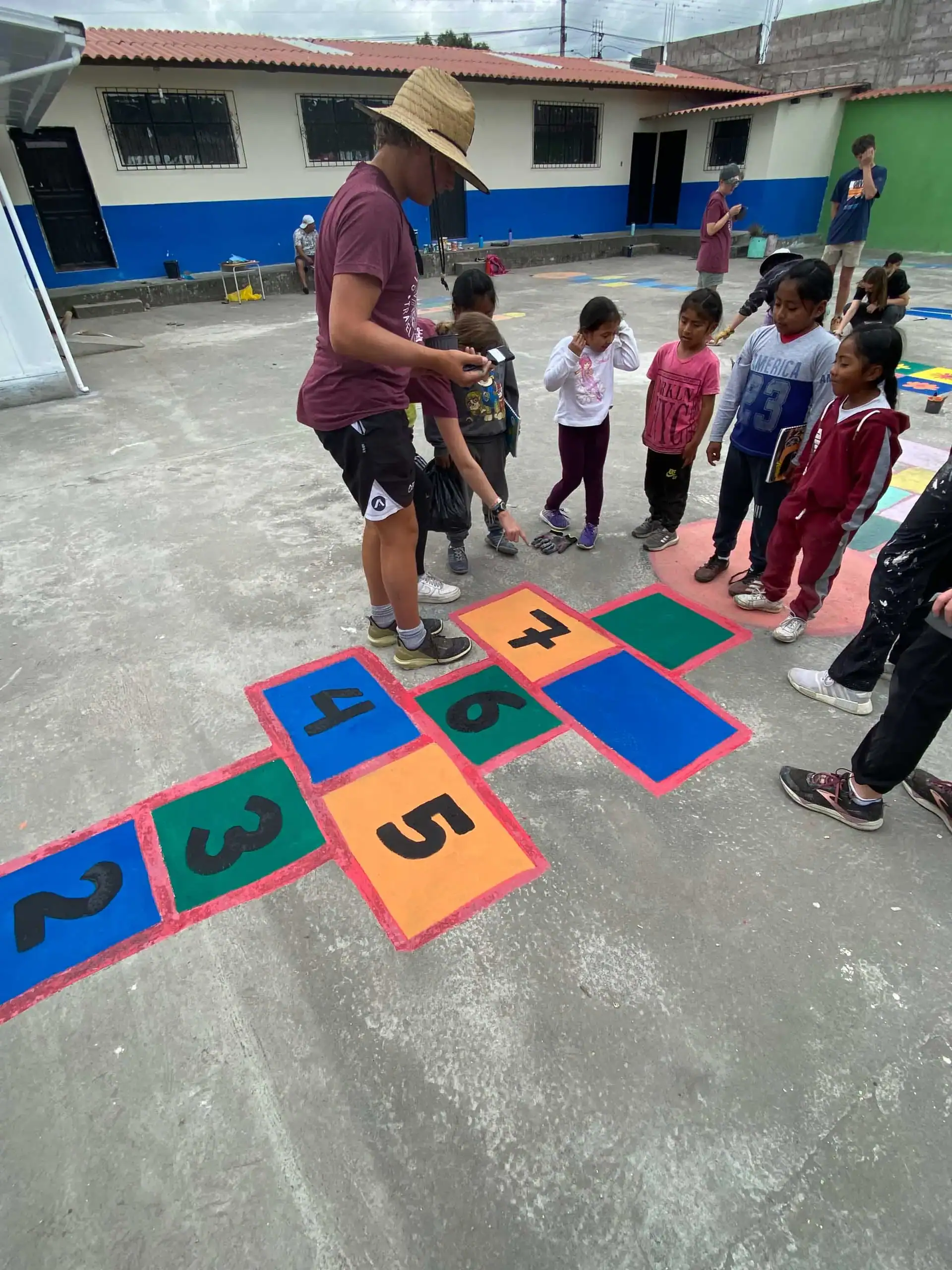 A group of children and an adult stand around a colorful hopscotch grid painted on a concrete surface in an outdoor area, similar to activities offered by teen programs that work with kids.