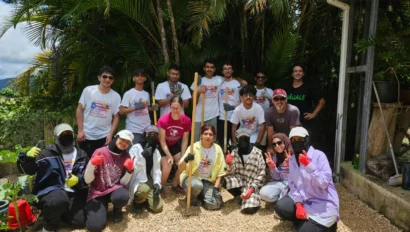 A group of people posing on a gravel path, some kneeling, some standing, with trees in the background. They are wearing casual clothing and gloves.