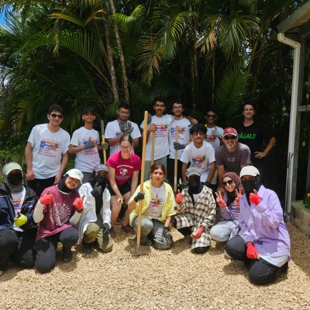 A group of people posing on a gravel path, some kneeling, some standing, with trees in the background. They are wearing casual clothing and gloves.