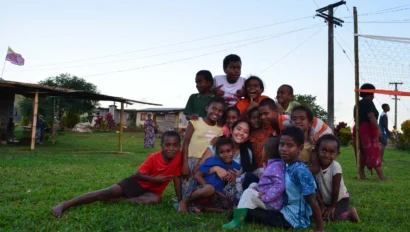 A group of children and an adult pose together on a grassy field near a volleyball net and utility poles, with a few people and houses in the background.
