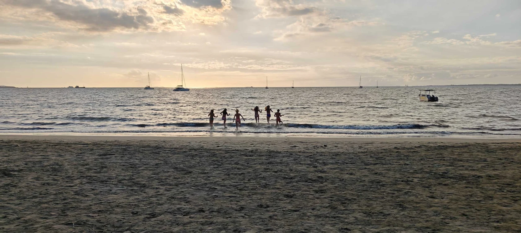 Silhouettes of people jumping in waves at a sandy beach during sunset, with sailboats in the background.
