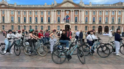 A group of people on bicycles gather in front of a historic building with a clock and flags on a clear day.