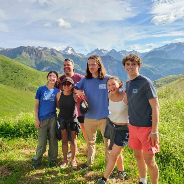 A group of five people smile and pose together on a grassy hillside, with a mountain range in the background under a clear blue sky.