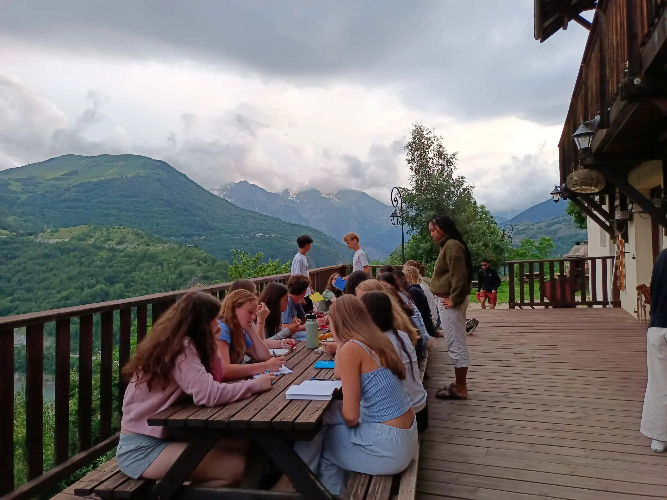 A group of people sit at wooden tables on a deck overlooking green mountains, brainstorming for their travel writing program, while others stand nearby under a cloudy sky.