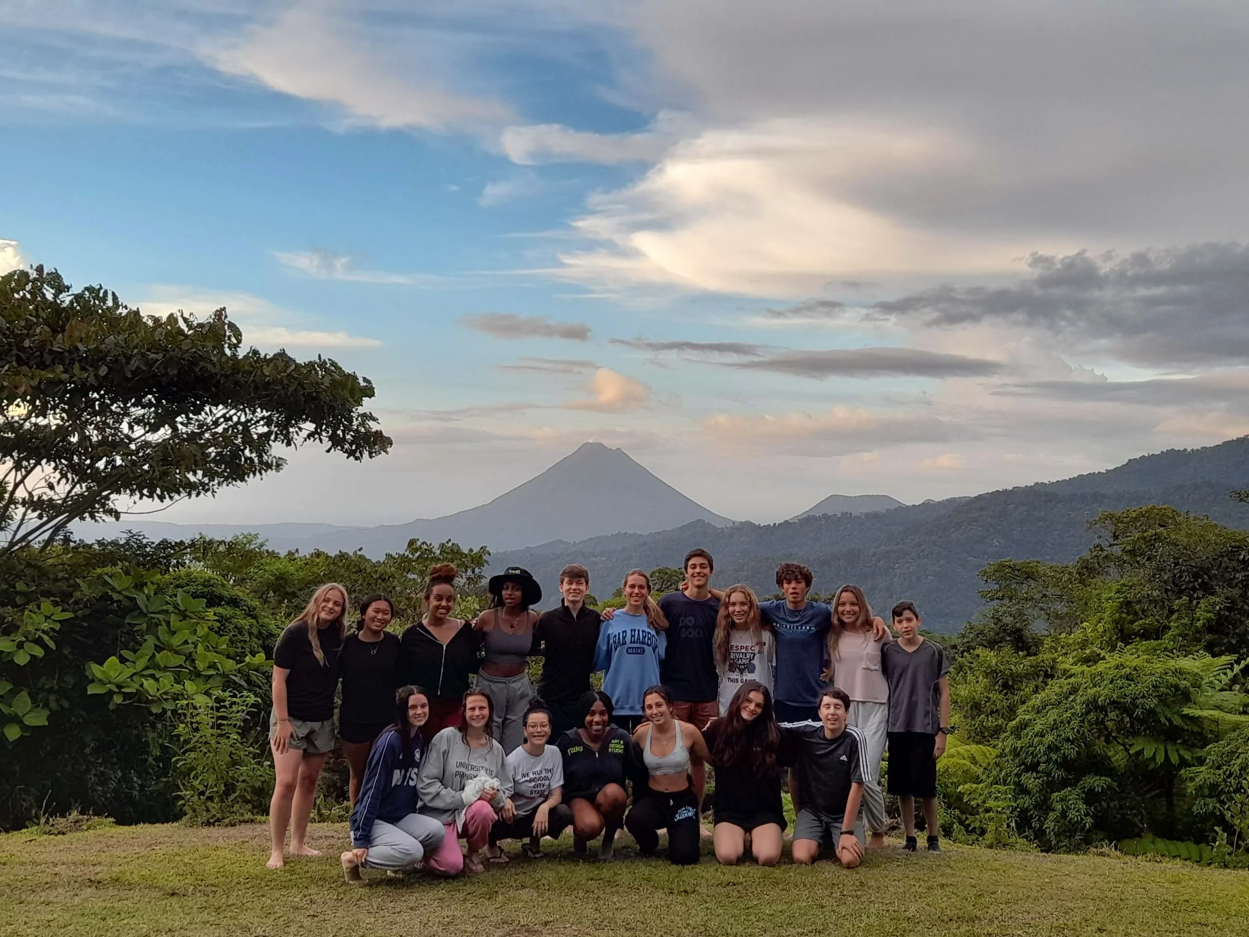 A group of people posing on a grassy area with a distant volcano and cloudy sky in the background.