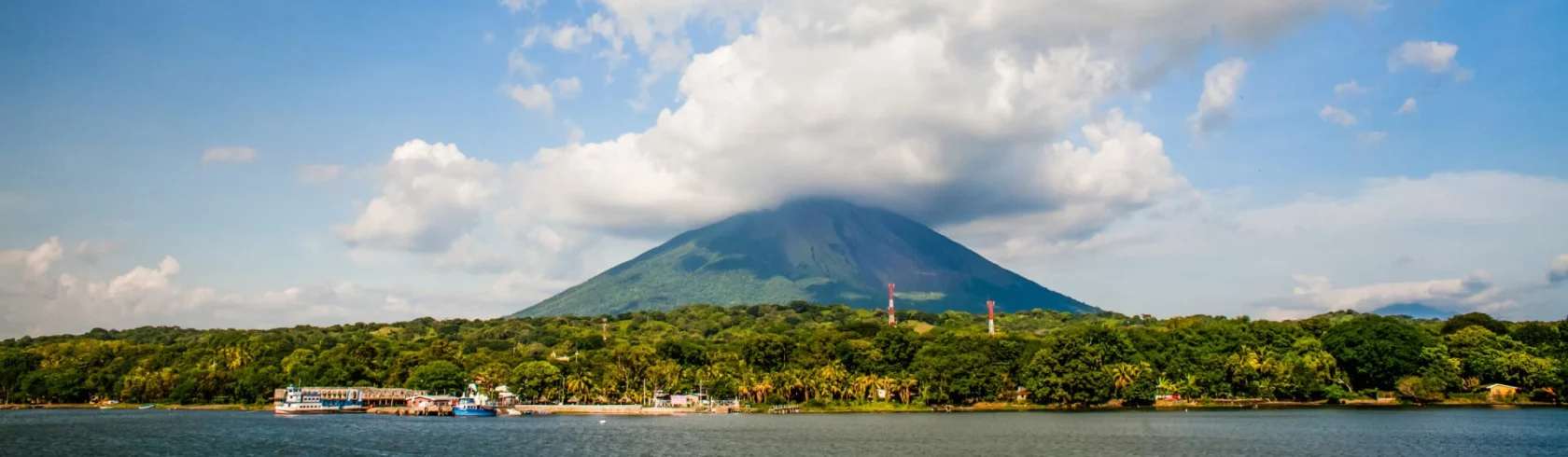 A lush green island with a volcano partially covered by clouds, surrounded by water. Boats are docked along the shore.
