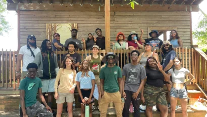 A diverse group of people posing together on wooden steps in front of a rustic building, with trees visible in the background. They appear relaxed and are casually dressed.